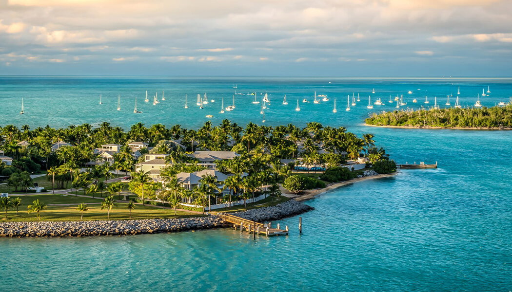 Aerial view of a lush tropical island resort with white buildings, palm trees, and a marina filled with sailboats, set against a turquoise ocean under a cloudy sky.