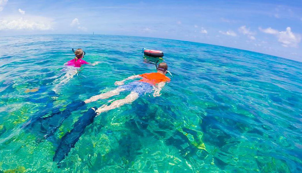Two people snorkel in crystal-clear turquoise ocean water, wearing masks and fins. One person in an orange shirt and blue shorts swims ahead, while another in a pink shirt follows. A red and black buoy floats nearby.
