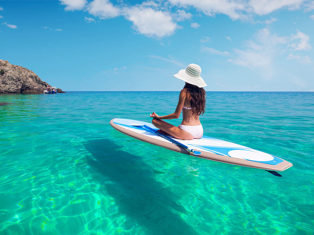 Woman in a white sun hat and bikini meditating on a paddleboard in crystal-clear turquoise water, with a rocky island and blue sky in the background.