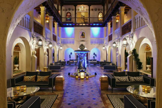 Luxurious hotel lobby with a central fountain, ornate architecture, plush seating, and ambient blue lighting.
