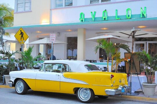 A vintage two-tone yellow and white convertible classic car parked in front of the Avalon Hotel, featuring palm trees and outdoor seating.