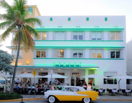 A classic yellow and white convertible parked in front of the Avalon hotel, a white Art Deco building with teal accents and neon signage, on a sunny day with outdoor diners.