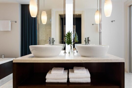 Modern hotel bathroom with a double vanity, featuring two white vessel sinks, polished chrome faucets, and a marble countertop. Pendant lights hang above the sinks, and neatly folded white towels are stacked below.
