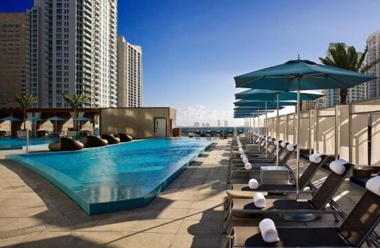 Luxury resort pool deck with blue lounge chairs and umbrellas, overlooking the ocean and city skyline. Modern high-rise buildings frame the tropical oasis.
