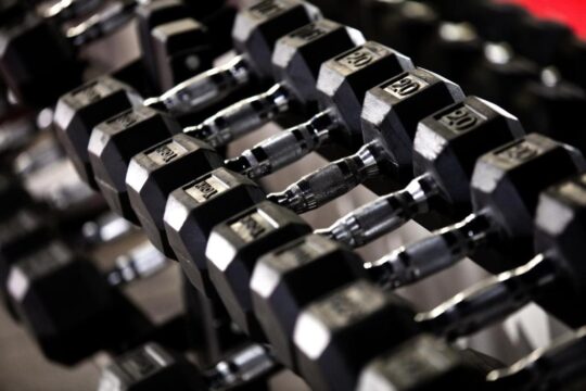 Close-up of a rack of black hex dumbbells, with weights ranging from 20 to 30 lbs, showcasing their textured grip and hexagonal shape.