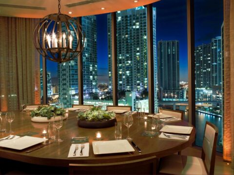 Elegant dining room with a large round table set for a meal, featuring floral centerpieces and a dramatic spherical chandelier against a backdrop of a glittering city skyline at night.