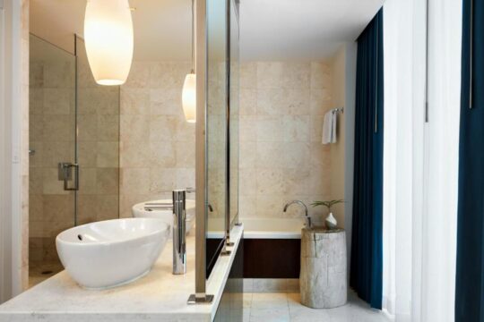 Modern bathroom with a white vessel sink on a marble countertop, a glass-enclosed shower, and a unique log-style vanity next to a bathtub, all set against textured beige tile walls and dramatic navy and white curtains.