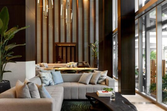 Luxury hotel lobby with a modern sectional sofa, natural wood accents, and large windows offering views of lush greenery. Contemporary interior design creates a serene and inviting atmosphere.