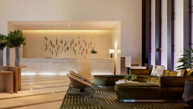 Luxurious hotel lobby with modern green velvet sectional sofa, wicker chairs, and a striking wall art installation above marble reception desks.