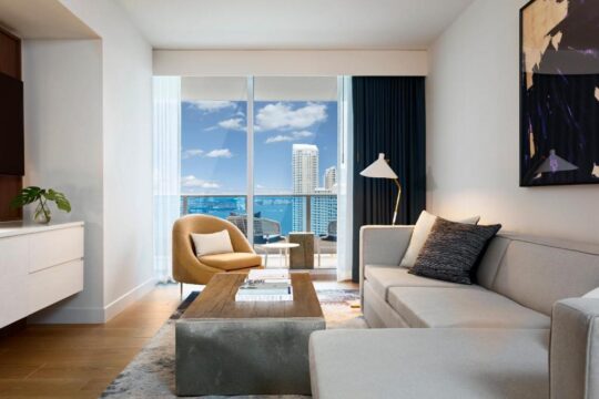 Modern hotel living room with large windows overlooking a city skyline and ocean, featuring a comfortable sofa, armchair, coffee table, and lamp.