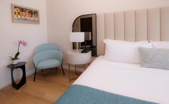 A serene hotel bedroom featuring a plush beige headboard, a crisp white bed with a teal throw blanket, a light blue armchair, and a small orchid on a black side table.
