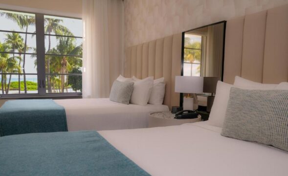 Two beds with teal and white linens in a bright hotel room with a large window overlooking palm trees and the ocean.