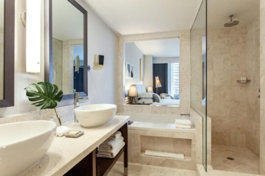 Luxurious hotel bathroom with dual vessel sinks, a deep soaking tub, and a walk-in shower with travertine tile, reflecting a modern spa-like ambiance.