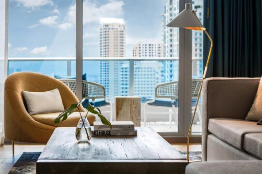 Modern living room with a mustard yellow armchair, a rustic wooden coffee table with books and a plant, and a floor lamp, overlooking a city skyline from a balcony.