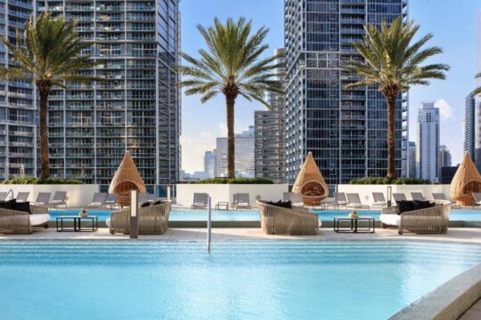 Luxury high-rise apartment building rooftop pool with palm trees, cabanas, and modern lounge seating, offering city views.