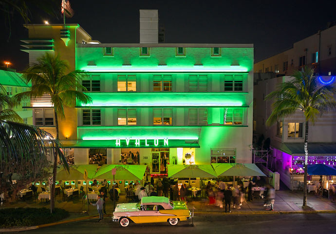 Avalon Hotel, Ocean Drive, Miami Beach at night — green-lit Art Deco facade with "AVALON" signage, sidewalk cafe seating, and a vintage pink-and-yellow classic car parked out front.