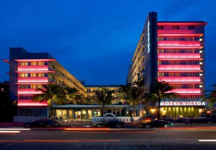 Hotel Victor, Ocean Drive, Miami Beach at dusk — pink-lit Art Deco facade with "HOTEL VICTOR" signage, palm-lined street, long-exposure car light trails in the foreground.