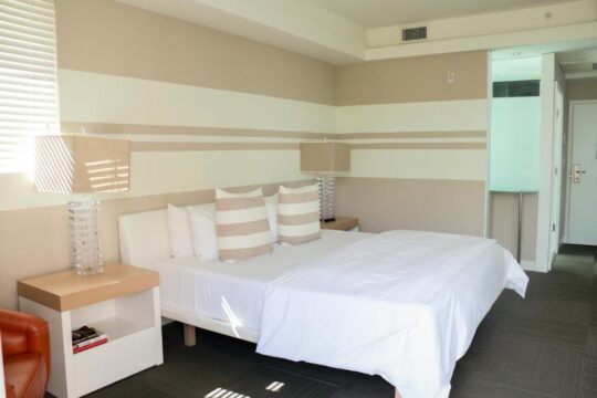 A modern hotel bedroom with a king-size bed, white linens, and striped accent pillows. A bedside table with a lamp and books sits next to the bed, with a striped accent wall behind.