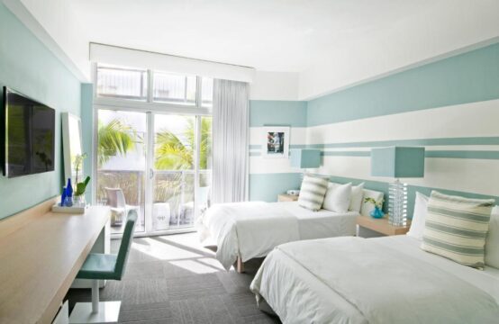 Bright and airy hotel room with two queen beds, featuring coastal-inspired decor with light blue and white striped walls, a large window with balcony access, a desk with a TV, and tropical palm trees visible outside.