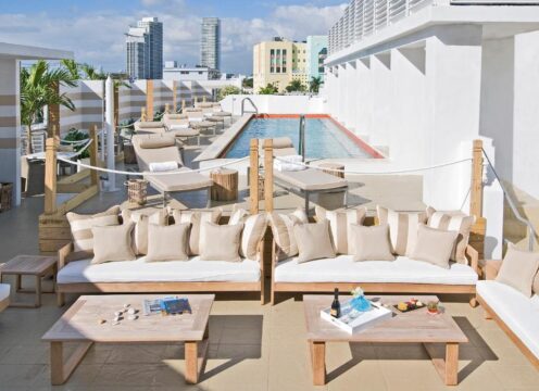 Rooftop lounge with white sofas, wooden tables, and a swimming pool, overlooking a city skyline and palm trees.