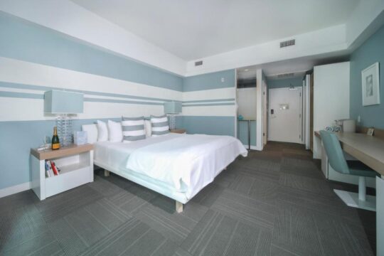 A modern hotel room with a king-size bed, blue and white striped walls, and a desk with a chair. The room has a minimalist design and a serene atmosphere.