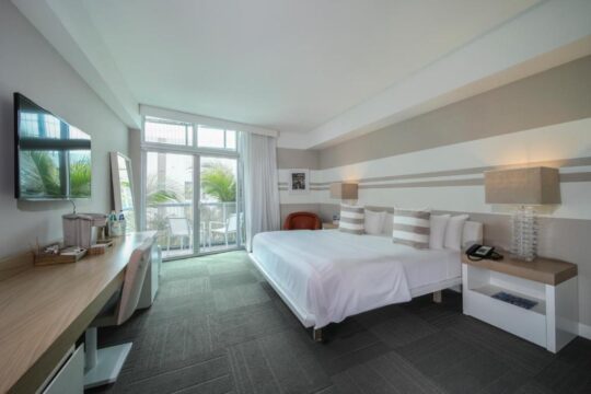 Modern hotel room with a king-size bed, striped pillows, and a balcony view of palm trees. A desk with a TV and amenities sits to the left.