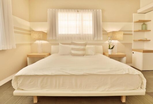 A serene bedroom with a large white bed, flanked by two bedside tables with lamps, a window with sheer curtains, and built-in shelving on the right.