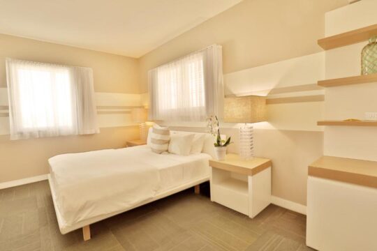A serene bedroom with a queen-size bed dressed in crisp white linens, flanked by a light wood nightstand with a textured lamp. The room features a soothing color palette of cream and beige stripes on the walls, with two windows letting in soft natural light.