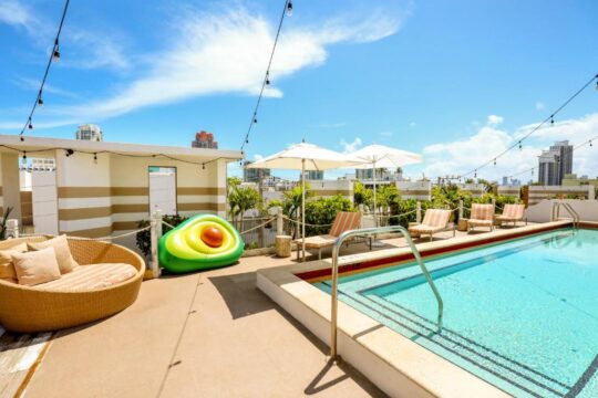 A vibrant poolside scene on a rooftop, featuring a large avocado float, lounge chairs, and umbrellas under a bright blue sky with string lights.