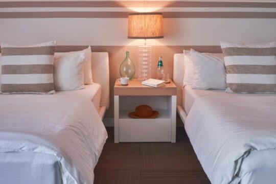 Two neatly made twin beds with white linens and striped pillows flank a bedside table featuring a lamp, a decorative vase, and a hat, creating a cozy hotel room ambiance.