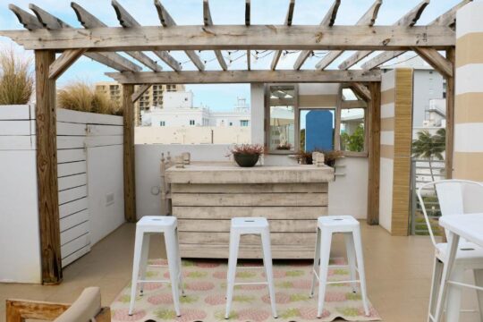 A rustic wooden pergola shades a rooftop bar area with a white wooden bar, three white stools, and a pineapple-patterned rug, overlooking a coastal city skyline.