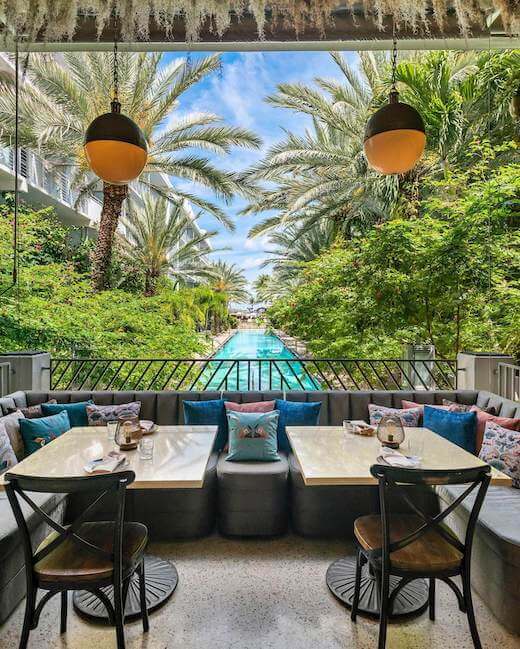 Elegant outdoor dining area with plush seating, tropical palm trees, and a sparkling blue pool in the background. Two tables are set for a meal, with modern pendant lights overhead.