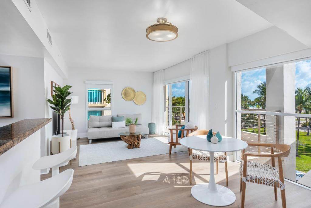 Bright, coastal-style suite at the Bentley Hotel South Beach — light wood floors, white walls, round marble dining table with woven-back chairs, ocean/palm view through floor-to-ceiling windows.