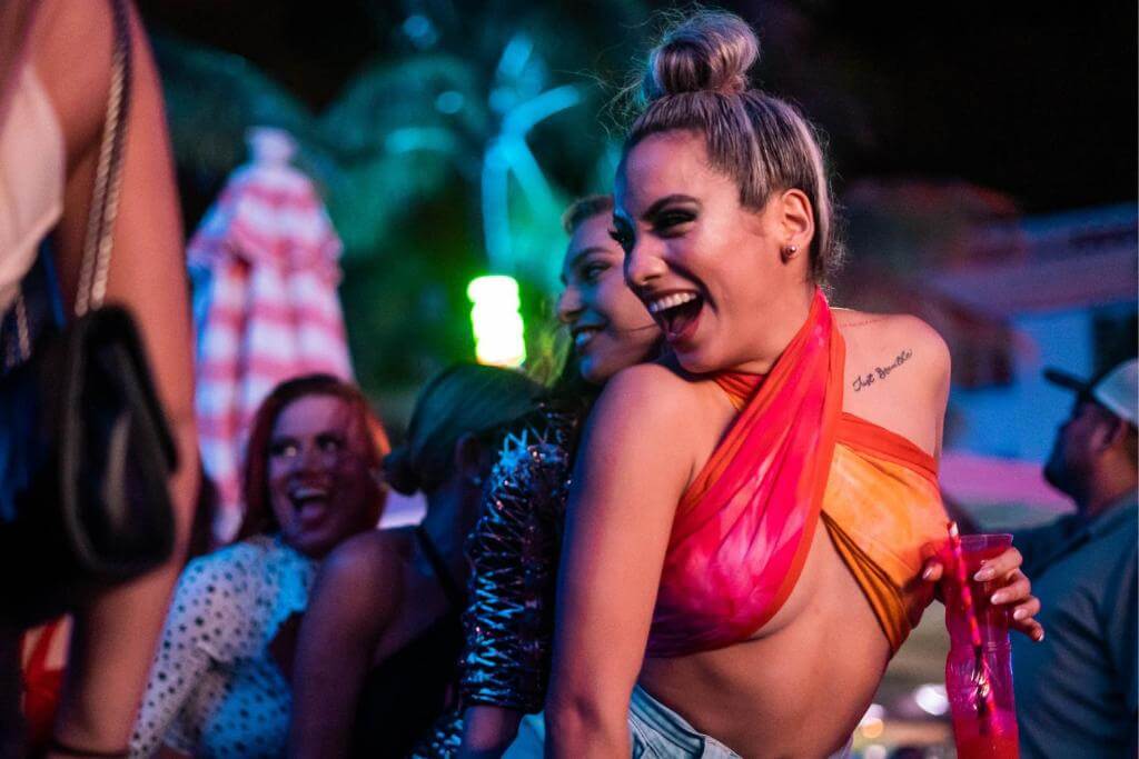 Nightlife scene at the Clevelander, Ocean Drive — laughing woman in a pink/orange wrap top in the foreground, group of friends behind her, colorful neon-lit poolside atmosphere.