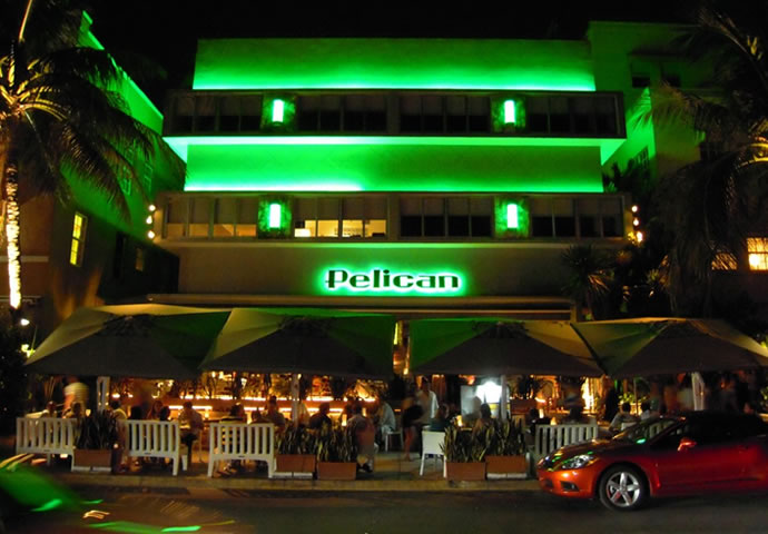 Pelican Hotel, Ocean Drive, Miami Beach at night — building lit in bold green, "Pelican" signage, ground-floor outdoor dining under umbrellas, palm trees framing the facade.