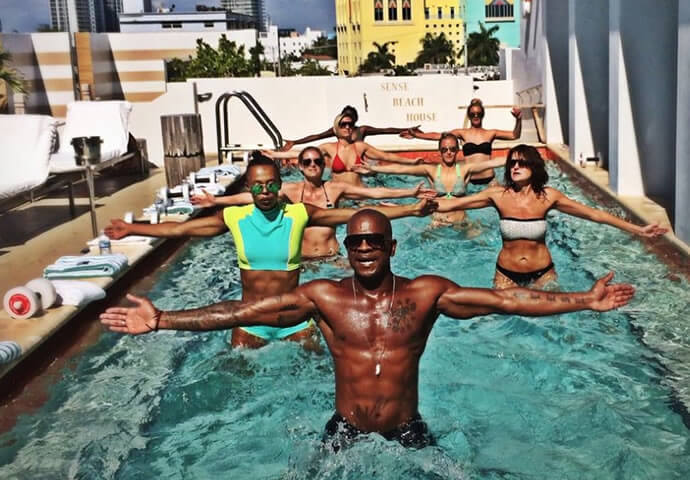 Rooftop pool group aqua-fitness class at Sense Beach House, Miami Beach — instructor leading arms-out pose, guests in swimwear, colorful Ocean Drive-style facades in the background.