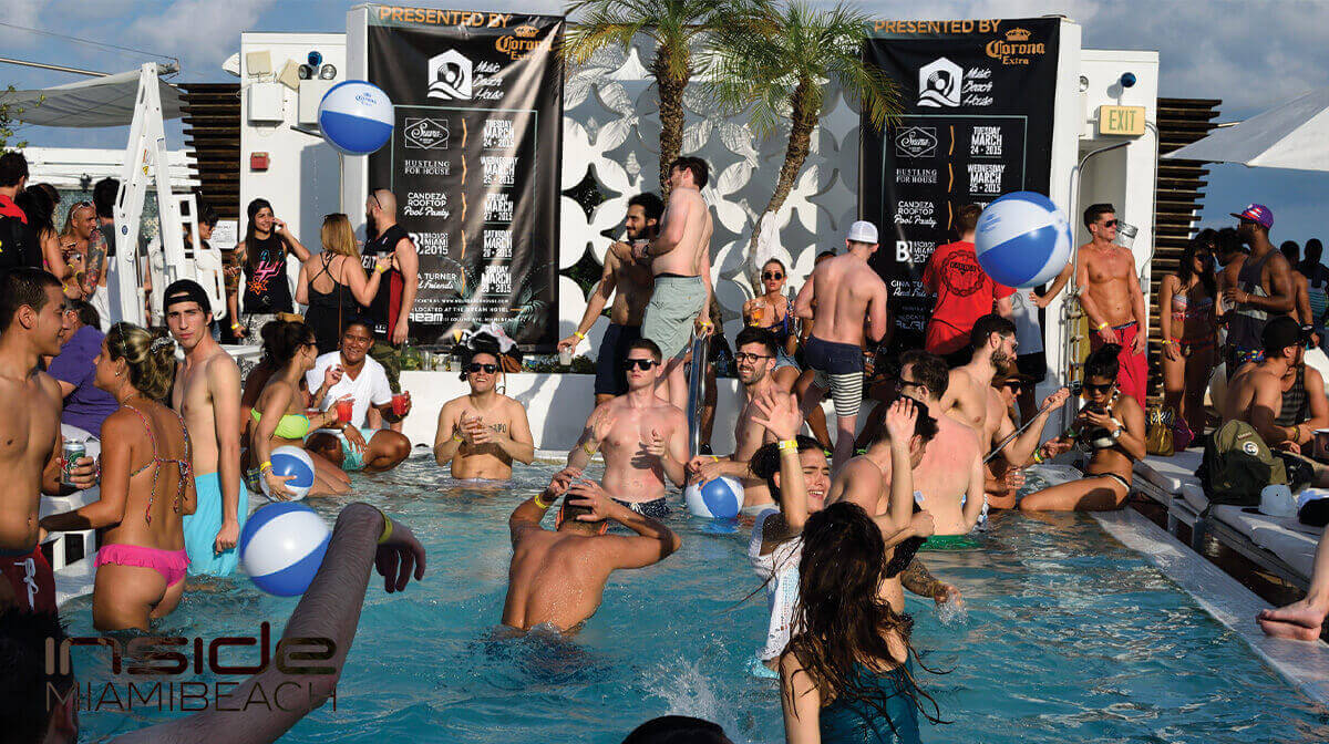 A lively pool party with people swimming, playing with beach balls, and socializing in and around the pool on a sunny Miami Beach day. Banners and palm trees are in the background.