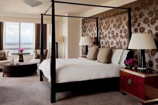 Luxurious hotel room with a four-poster bed, floral wallpaper, and a city view. Two bedside lamps illuminate the plush pillows and white bedding.
