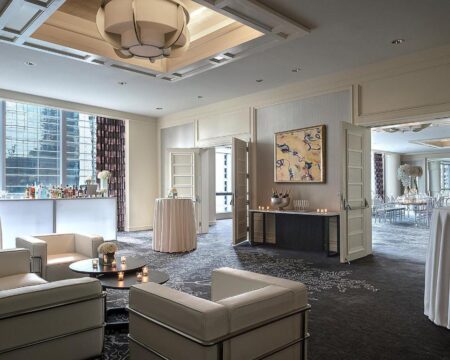 Elegant lounge area with white leather sofas, a bar stocked with bottles, and a high-top table with draped cloth, set against a backdrop of large windows and a sophisticated event space.