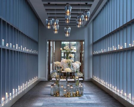 Elegant hotel hallway with modern glass pendant lights and a central floral arrangement, illuminated by rows of glowing candles on both sides.