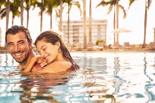 A joyful couple embraces in a sun-drenched swimming pool, with palm trees and a resort building in the blurred background. The man smiles broadly, holding the woman, who rests her head on his shoulder with her eyes closed.