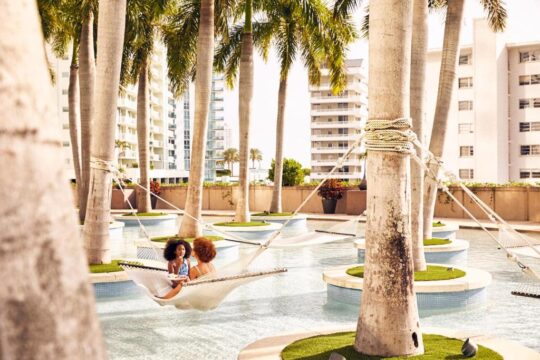 Two women relax in a hammock suspended over a clear blue swimming pool, surrounded by lush palm trees and modern white buildings, evoking a serene tropical resort atmosphere.