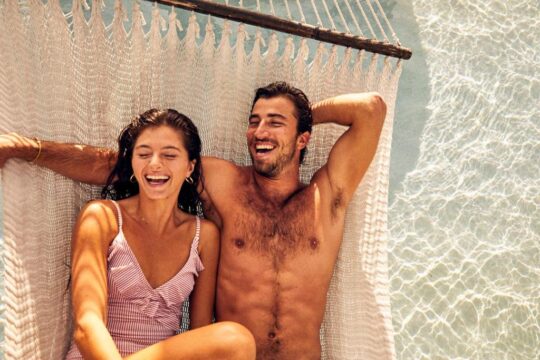 A joyful couple laughs while relaxing in a hammock suspended over crystal-clear turquoise water, embodying a perfect vacation getaway.