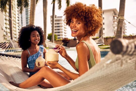 A Black mother with voluminous curly red hair and her young daughter with dark curly hair relax in a hammock over a pool, smiling and drinking from coconuts. Palm trees and a modern building are in the background.