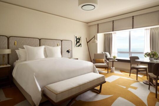 Luxurious hotel room with a king-size bed, ocean view, and modern decor. The room features a large window with a seating area, a round dining table, and a plush rug.