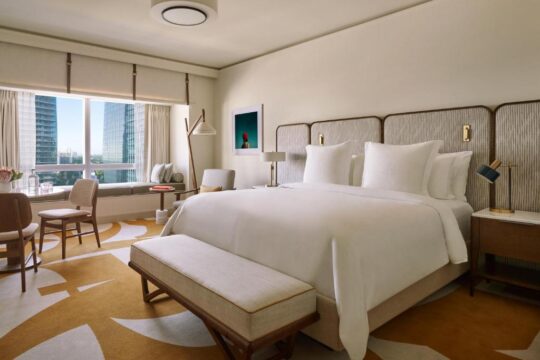 Luxurious hotel suite bedroom with a king-size bed, white linens, modern decor, city view window, and a stylish bench.
