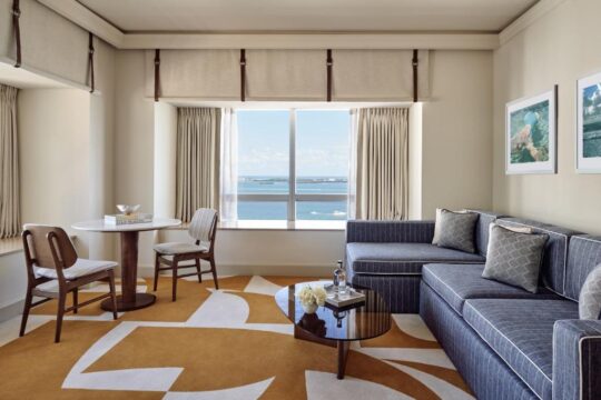 A luxurious hotel suite living room with a blue sectional sofa, a round dining table with two chairs, and a large window overlooking the ocean. The room features a striking abstract rug in warm ochre and white.