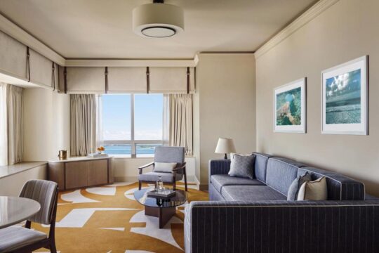 A stylish hotel suite living room with a blue sofa, armchair, and coffee table, featuring a large window with ocean views and modern artwork.