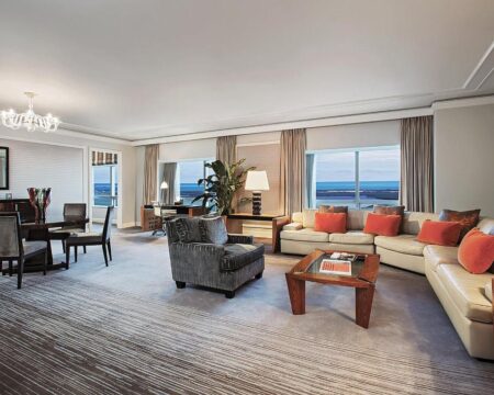 Luxurious hotel suite living room with a large sectional sofa, armchairs, coffee table, and dining area, featuring large windows with ocean views.
