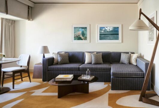 A modern hotel suite living area features a grey sectional sofa with pinstripes and decorative pillows, a dark coffee table with a book titled "Four Seasons," and a large abstract yellow and white rug.